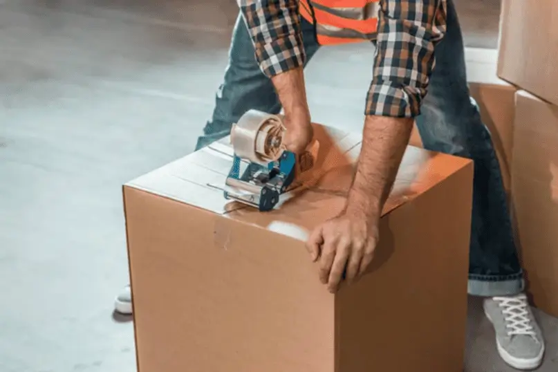 Trabajador sellando una caja de cartón con cinta adhesiva en un almacén, parte del proceso logístico relacionado con qué es carga consolidada y preparación de paquetes para envíos compartidos.